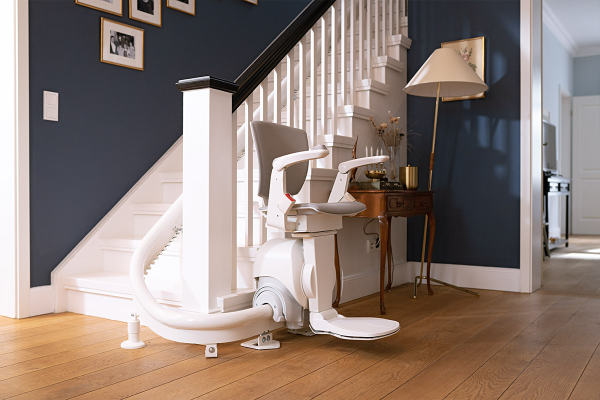 Moderner Treppenlift ist an einer Treppe mit marineblauer Wand und weißem Geländer installiert der ist betriebsbereit und befindet sich am Fuß der Treppe der Raum verfügt über einen Holzboden einen Tisch und eine Stehlampe an der Wand hängen Rahmen
