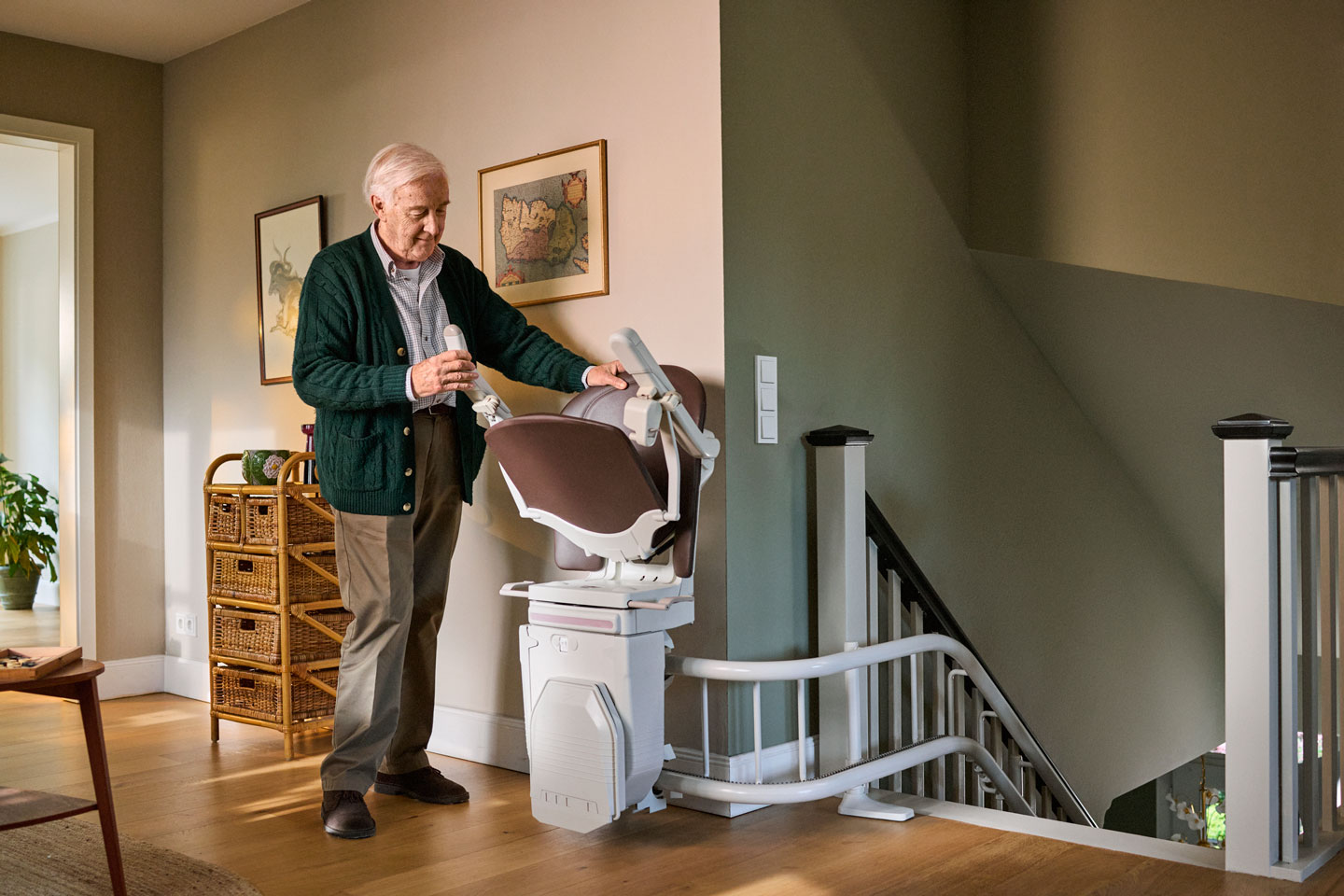 Älterer Mann steht neben einem Treppenlift in einem Wohnhaus er hält einen weißen Gegenstand in der Hand und berührt dabei den Sitz des Treppenlifts der sich oben auf einer Treppe befindet. Der Raum ist warm beleuchtet im Hintergrund sind gerahmte Bilder und ein Korbschrank zu sehen