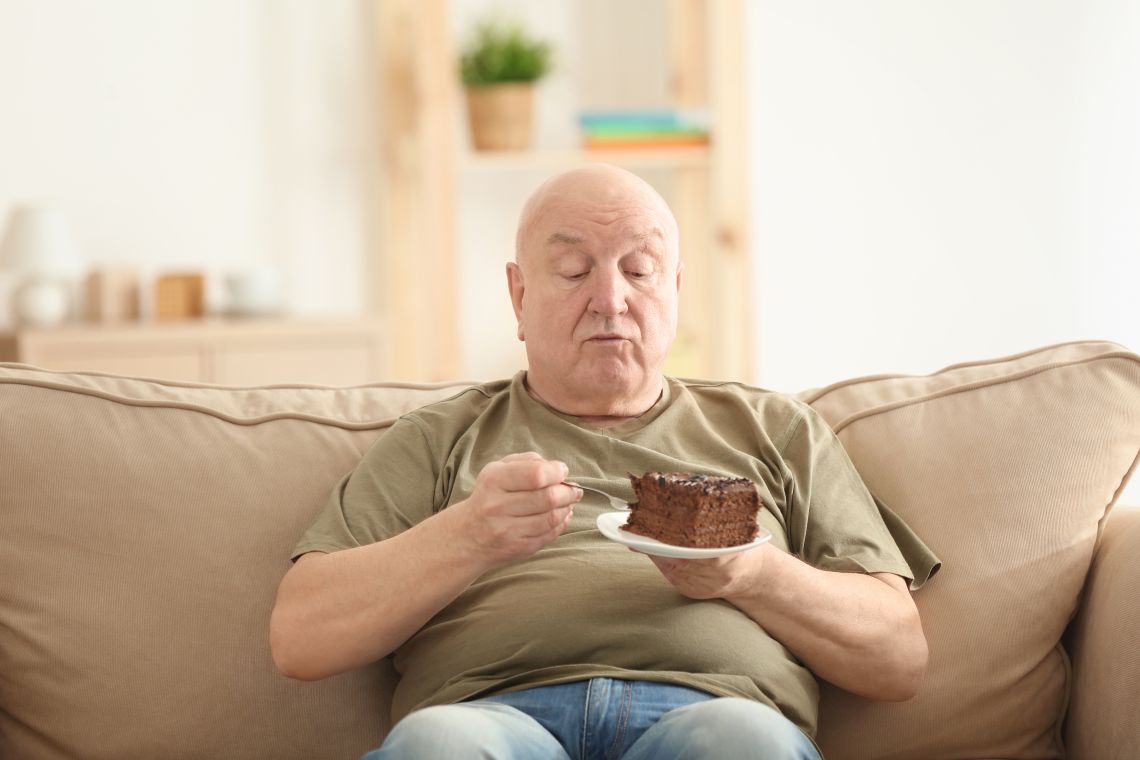 Ein älterer Mann mit Übergewicht hält einen Teller mit einem Stück Schokoladenkuchen in der Hand