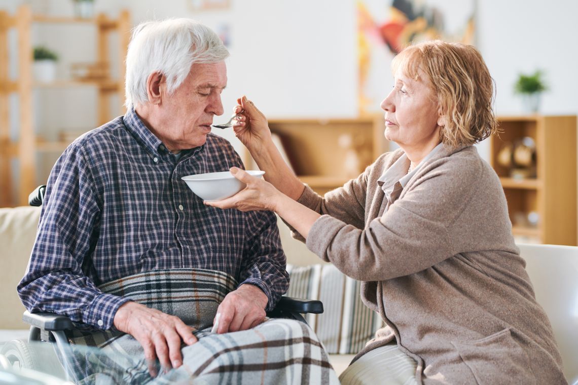 Eine ältere Frau füttert ihren Mann im Rollstuhl mit Suppe