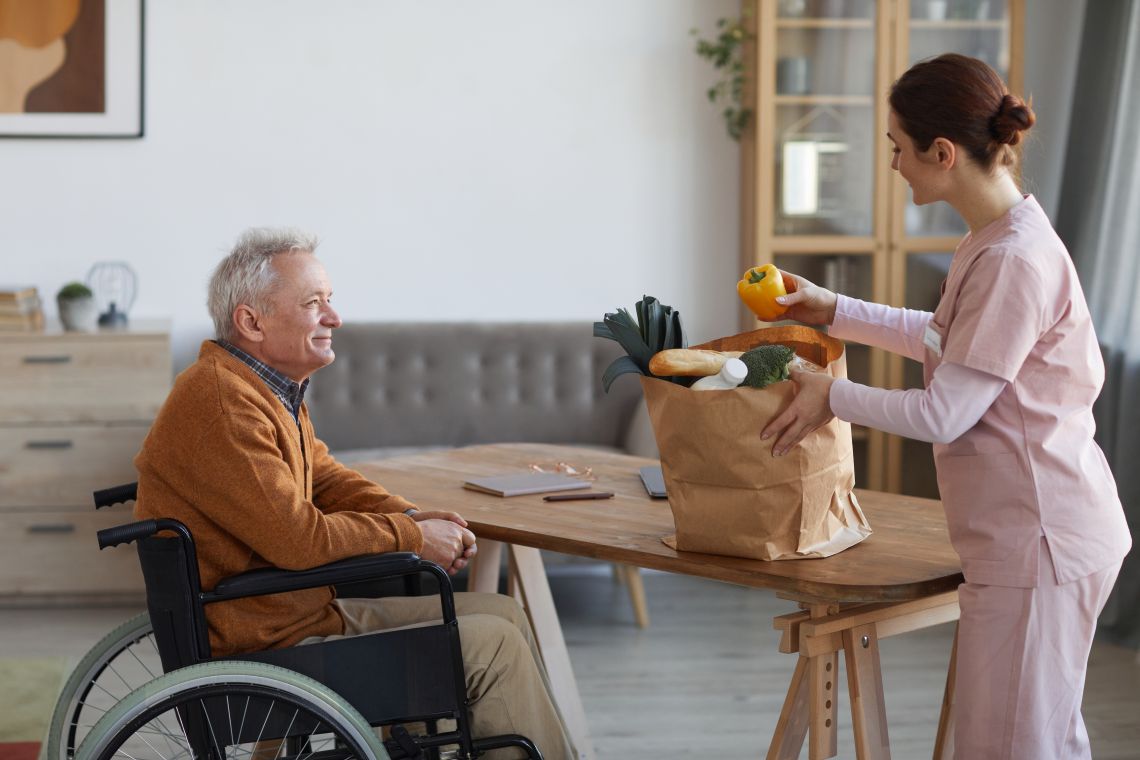 Eine Pflegerin packt den Einkauf für einen Senior im Rollstuhl aus