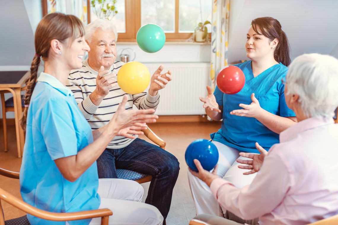 Zwei Senioren machen gemeinsam mit zwei Physiotherapeutinnen Gymnastik im Sitzen mit einem Ball