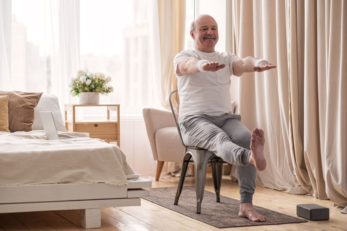 Ein älterer Mann macht eine Gymnastikübung auf einem Stuhl in seinem Schlafzimmer.